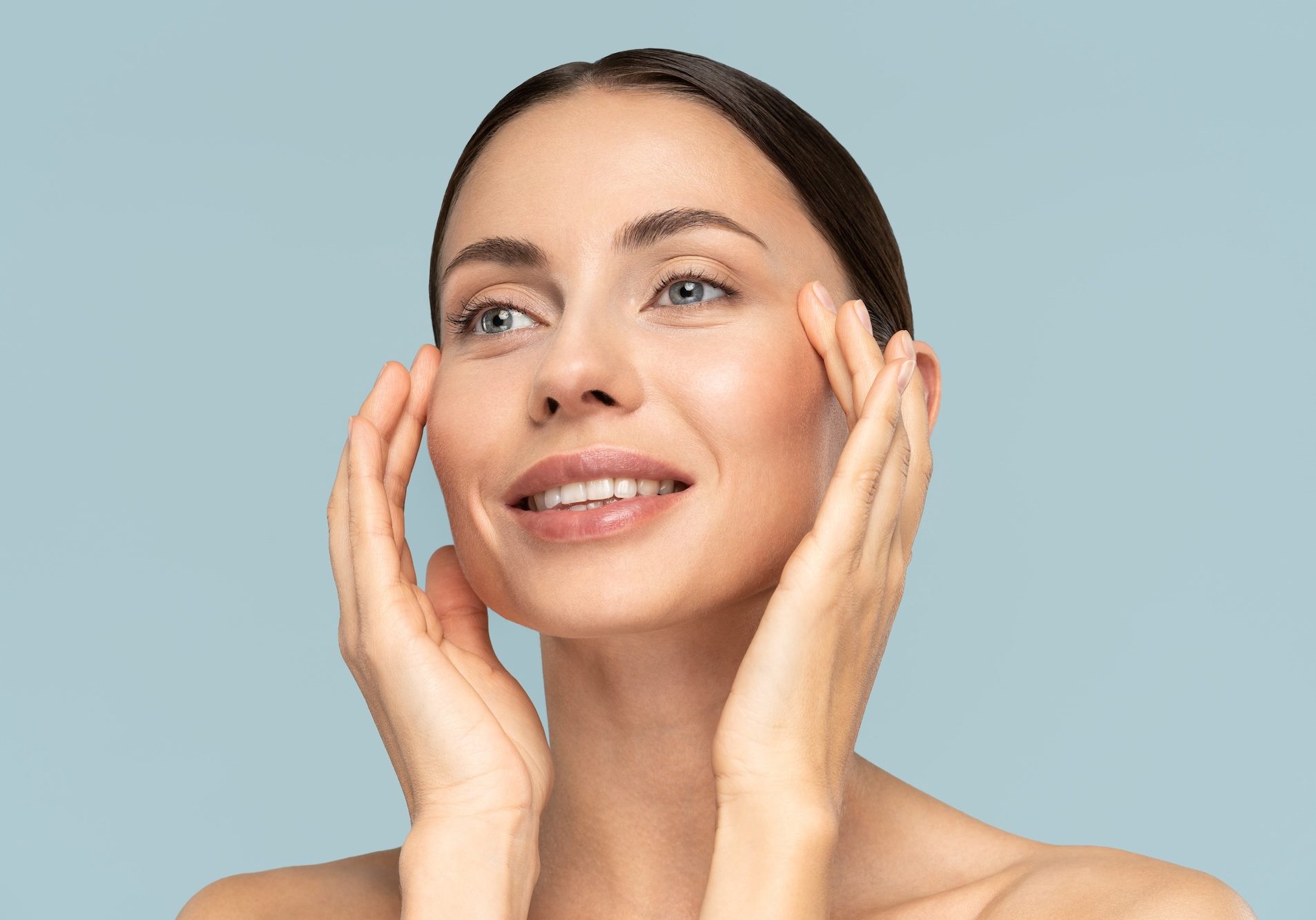 Studio portrait of young woman with natural makeup, combed hair, touching well-groomed pure skin on face, isolated on blue background. Beauty, facelift, anti-aging cosmetic procedure. Banner. Studio portrait of young woman with natural makeup, combed hair, touching well-groomed pure skin on face, isolated on blue background. Beauty, facelift, anti-aging cosmetic procedure. Banner.
